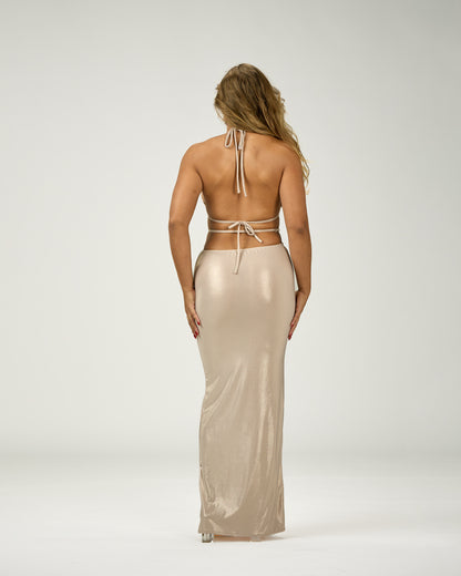 A woman viewed from the back wearing a shimmering champagne-beige maxi dress with a tie-strap halter-style back and a fitted silhouette, photographed against a plain light gray studio background.